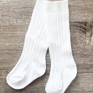 Può includere: Un paio di calzini bianchi a coste. I calzini sono alti fino al ginocchio, con una texture a coste verticali. Realizzati in un materiale morbido e opaco, sono progettati per il comfort. Esposti su una superficie di legno.