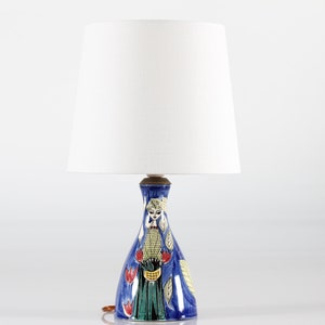 以下が含まれることがあります： 白い布製のシェードが付いた青と白のセラミック製のテーブルランプ。ランプのベースには、赤い花と黄色の葉がついた緑のドレスを着た女性の絵が描かれています。