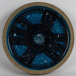 以下が含まれることがあります： 濃い青とティール色の釉薬を施した丸いセラミックプレート。中央には濃い青色のスターバーストのデザインがあります。縁は明るい茶色です。この装飾的なプレートは、食べ物を提供したり、装飾品として使用するのに適しています。
