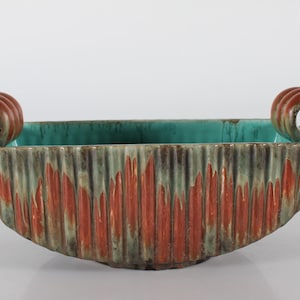 Puede incluir: Un cuenco de cerámica con dos asas. El cuenco es verde y marrón con una superficie texturizada. Las asas son marrones y tienen un diseño en espiral.