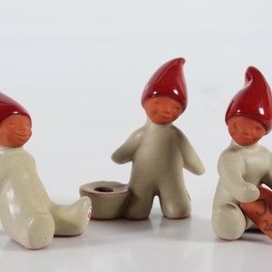 May include: Three ceramic figurines of gnomes wearing red hats. The gnome on the right is holding a heart-shaped ornament that says "Jul 2000".