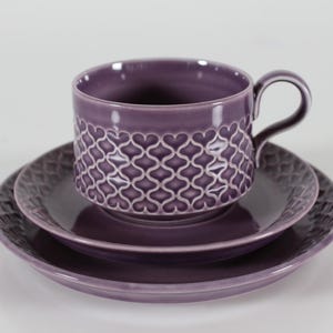 Puede incluir: Un juego de taza y platillo de cerámica morada. La taza tiene un asa y un patrón decorativo de formas entrelazadas. El juego incluye dos platillos a juego. El color es un morado profundo y apagado.