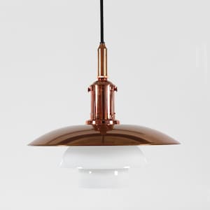 Puede incluir: Una moderna lámpara colgante con una pantalla y un vástago de color cobre, y un difusor de vidrio blanco en capas. La luz cuelga de un cable negro sobre un fondo blanco. El diseño del accesorio es elegante y minimalista.