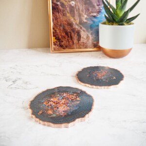 May include: Two decorative coasters with a dark blue and copper design. The coasters have a natural, irregular edge and a copper rim. A framed landscape print and a potted succulent are in the background.