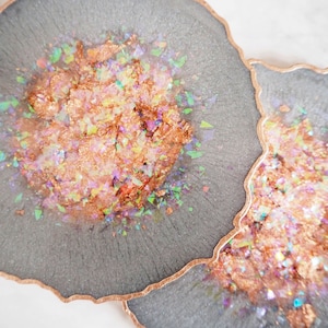 May include: Two grey resin coasters with a copper-coloured edge. The coasters have a glittery, iridescent finish with pink, green, and gold glitter.