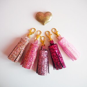 May include: Five sparkly pink and gold tassel keychains with gold key rings. The tassels are different shades of pink, from light pink to dark pink.