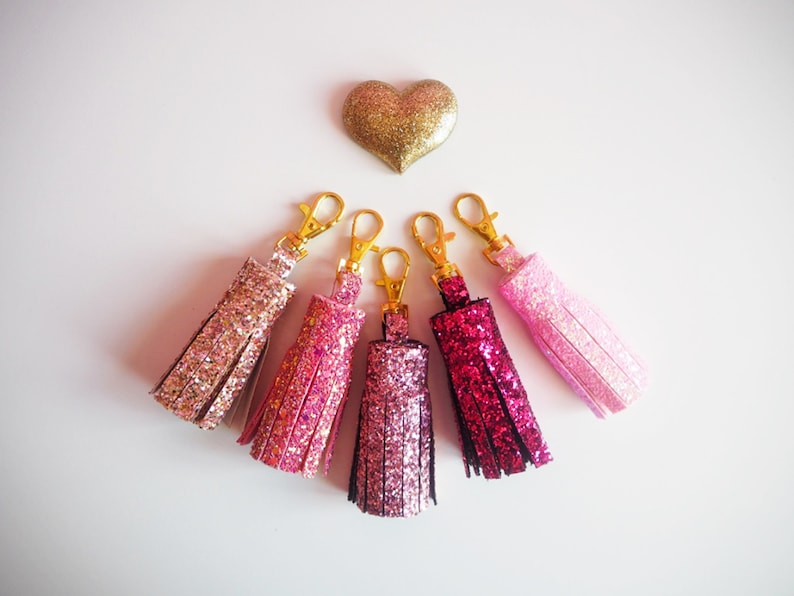 May include: Five sparkly pink and gold tassel keychains with gold key rings. The tassels are different shades of pink, from light pink to dark pink.