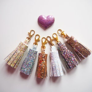 May include: Six sparkly keychains with gold clasps. The keychains are in various colours, including gold, silver, white, and pink. The keychains are arranged in a semi-circle on a white background.