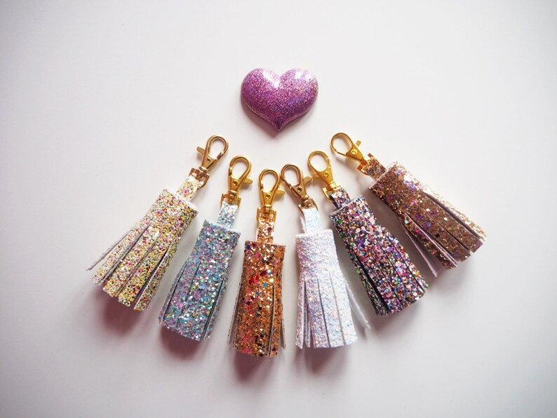 May include: Six sparkly keychains with gold clasps. The keychains are in various colours, including gold, silver, white, and pink. The keychains are arranged in a semi-circle on a white background.