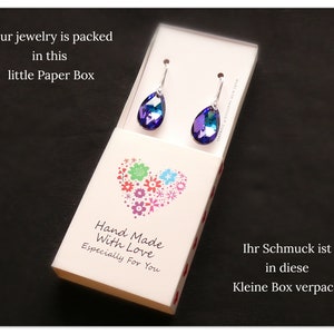 Pu&ograve; includere: Una piccola scatola bianca con un design a forma di cuore e il testo "Hand Made With Love Especially For You". La scatola contiene un paio di orecchini a forma di goccia con pietre iridescenti blu e viola.