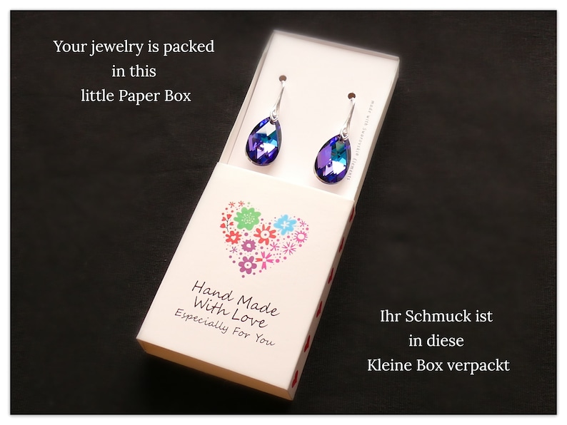 Pu&ograve; includere: Una piccola scatola bianca con un design a forma di cuore e il testo "Hand Made With Love Especially For You". La scatola contiene un paio di orecchini a forma di goccia con pietre iridescenti blu e viola.
