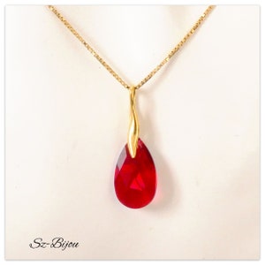 Peut inclure: Un collier doré avec un pendentif rouge en forme de goutte d'eau facettée. Le pendentif est suspendu à une fine chaîne dorée. Le bijou est présenté sur fond blanc. Le texte "Sz-Bijou" est dans le coin inférieur gauche.