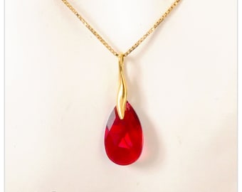 Pode incluir: Um colar dourado com um pingente vermelho facetado em forma de gota. O pingente é suspenso por uma delicada corrente dourada. A joia é exibida em um fundo branco. O texto "Sz-Bijou" está no canto inferior esquerdo.