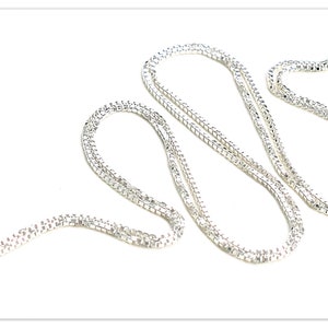 Op de afbeelding: Een zilverkleurige box chain ketting met een reeks onderling verbonden vierkante schakels die een gladde, doorlopende textuur creëren. De ketting is gerangschikt in een los, kronkelend patroon en bevat een ronde veerringsluiting aan één uiteinde.