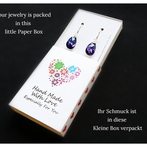 Pu&ograve; includere: Una scatola bianca con un design a forma di cuore e il testo "Hand Made With Love Especially For You". La scatola contiene un paio di orecchini a goccia di colore viola.