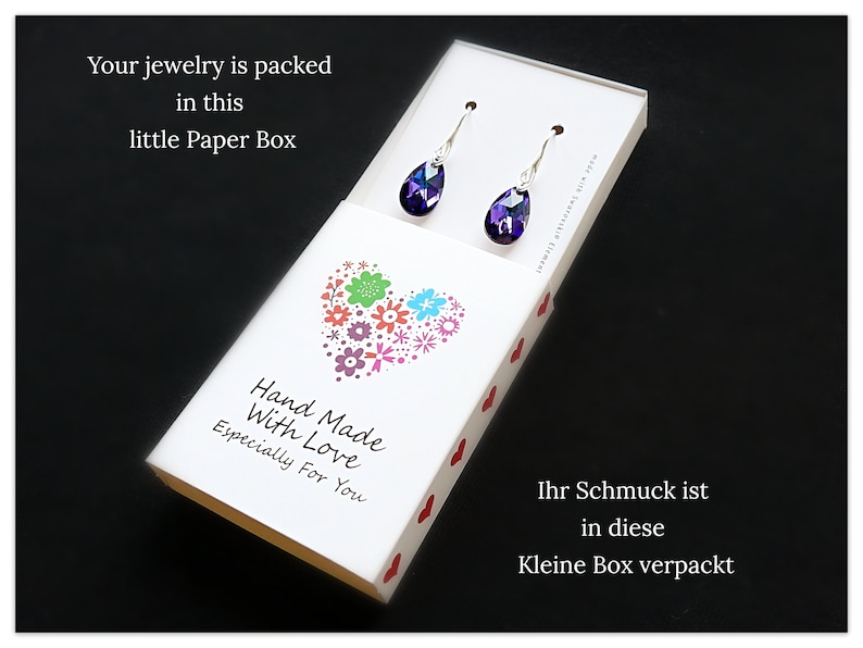 Pu&ograve; includere: Una scatola bianca con un design a forma di cuore e il testo "Hand Made With Love Especially For You". La scatola contiene un paio di orecchini a goccia di colore viola.