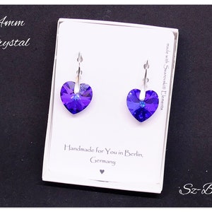 May include: A pair of silver heart-shaped earrings with purple crystal stones. The earrings are displayed on a white card with the text "14mm Crystal", "made with Swarovski Elements", "Handmade for You in Berlin, Germany", and "Sz-Bijou".