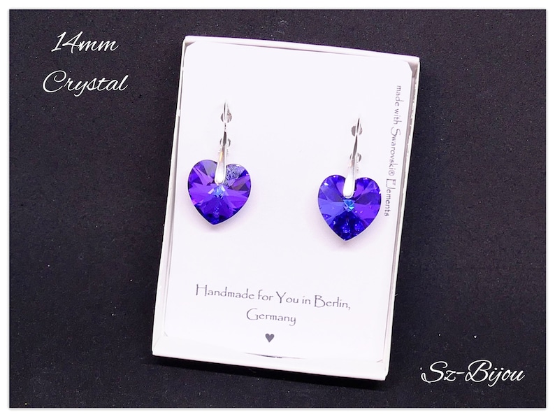 May include: A pair of silver heart-shaped earrings with purple crystal stones. The earrings are displayed on a white card with the text "14mm Crystal", "made with Swarovski Elements", "Handmade for You in Berlin, Germany", and "Sz-Bijou".