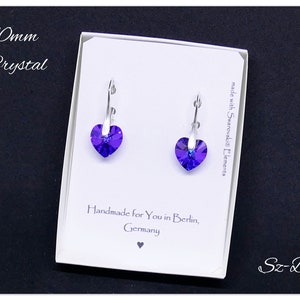 May include: A pair of silver heart-shaped earrings with purple crystal stones. The earrings are in a white box with the text "10mm Crystal", "made with Swarovski Elements", "Handmade for You in Berlin, Germany", and "Sz-Bijou".