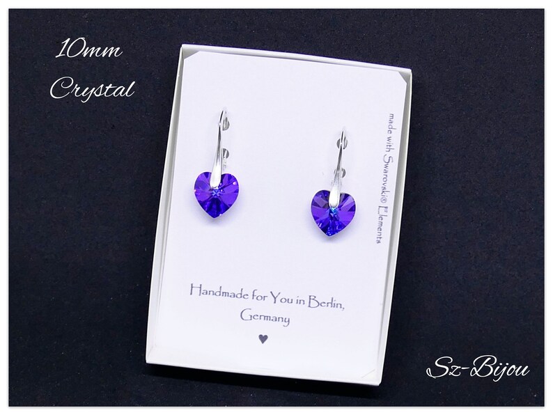 May include: A pair of silver heart-shaped earrings with purple crystal stones. The earrings are in a white box with the text "10mm Crystal", "made with Swarovski Elements", "Handmade for You in Berlin, Germany", and "Sz-Bijou".