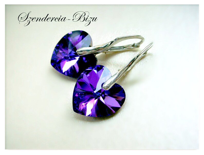 May include: A pair of silver heart-shaped earrings with purple crystal accents. The earrings have a hook closure.