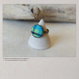 Puede incluir: Anillo con dise&ntilde;o de tela turquesa, azul y verde. El anillo tiene una banda de color bronce y un borde festoneado. Se muestra sobre un soporte c&oacute;nico blanco. El texto dice "BAGUES EN TISSUS MADRAS".