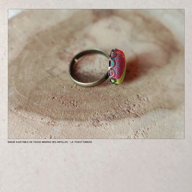 Puede incluir: Un anillo ajustable con un engaste rectangular de tela colorida. El anillo tiene una banda de color bronce y el engaste de tela muestra tonos rojos, amarillos y verdes. El anillo est&aacute; sobre una superficie de madera.