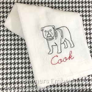 K&ouml;nnte beinhalten: Ein wei&szlig;es K&uuml;chentuch mit einer schwarzen Stickerei, die den Umriss einer Bulldogge im Profil zeigt. Unter dem Hund ist das Wort Cook in roter Schreibschrift gestickt. Das Tuch ist gefaltet und zeigt ein schlichtes, klares Design.