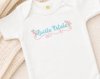 May include: A white short-sleeved baby bodysuit featuring the embroidered name Lucila Estela in teal cursive script, accented by pink floral motifs and decorative scrollwork. The garment includes a round neckline and three metal snap closures.