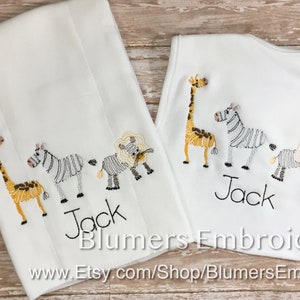 May include: A white burp cloth and a white baby bib, both featuring embroidered designs of a giraffe, a zebra, and a lion. The name Jack is embroidered in black thread below the animals on each item. The text Blumers Embroidery is visible.