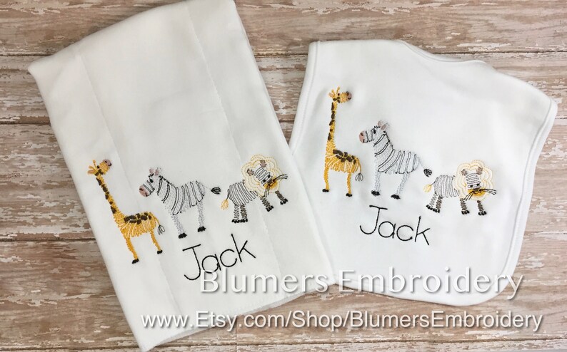 May include: A white burp cloth and a white baby bib, both featuring embroidered designs of a giraffe, a zebra, and a lion. The name Jack is embroidered in black thread below the animals on each item. The text Blumers Embroidery is visible.