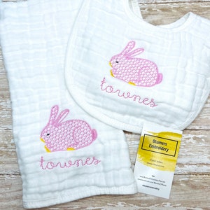 May include: A white burp cloth and bib with pink embroidered bunnies and the name "Towenes" in yellow thread. The bib is folded in half and the burp cloth is folded in thirds. A business card for Blumers Embroidery is visible in the bottom right corner.