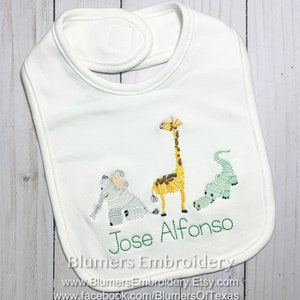 May include: A white baby bib featuring an embroidered design of a light blue elephant, a yellow giraffe, and a green alligator. The name Jose Alfonso is embroidered in green thread below the animals. The bib has a hook and loop closure at the neck.