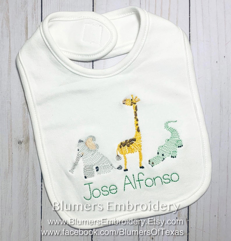 May include: A white baby bib featuring an embroidered design of a light blue elephant, a yellow giraffe, and a green alligator. The name Jose Alfonso is embroidered in green thread below the animals. The bib has a hook and loop closure at the neck.