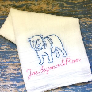K&ouml;nnte beinhalten: Ein wei&szlig;es Stoffhandtuch mit einer blauen Stickerei, die den Umriss einer Bulldogge zeigt. Unter dem Hund sind die Namen Joe, Seyma und Ron in rosa Schreibschrift gestickt. Das Handtuch hat eine weiche, strukturierte Oberfl&auml;che und ist gefaltet.