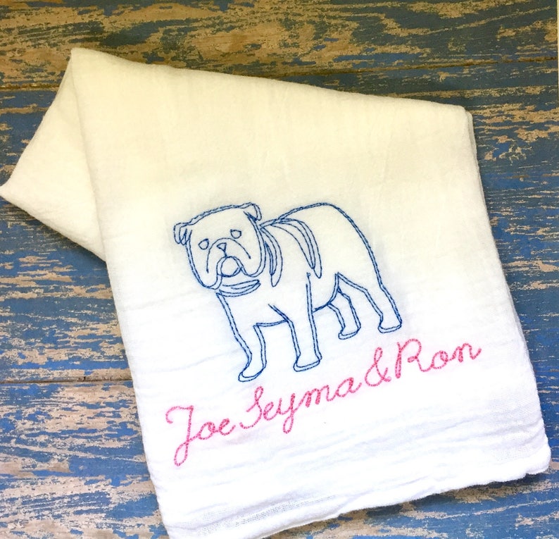 K&ouml;nnte beinhalten: Ein wei&szlig;es Stoffhandtuch mit einer blauen Stickerei, die den Umriss einer Bulldogge zeigt. Unter dem Hund sind die Namen Joe, Seyma und Ron in rosa Schreibschrift gestickt. Das Handtuch hat eine weiche, strukturierte Oberfl&auml;che und ist gefaltet.