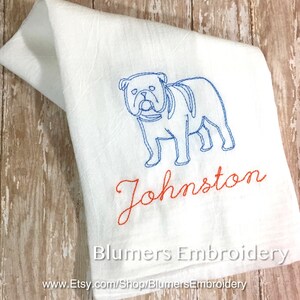 K&ouml;nnte beinhalten: Ein wei&szlig;es K&uuml;chentuch mit einer blauen Stickerei, die den Umriss einer Bulldogge zeigt. Unter dem Hund ist der Name Johnston in orangefarbener Schreibschrift gestickt. Der Text Blumers Embroidery und eine URL sind unten im Bild zu sehen.