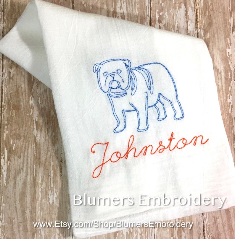 K&ouml;nnte beinhalten: Ein wei&szlig;es K&uuml;chentuch mit einer blauen Stickerei, die den Umriss einer Bulldogge zeigt. Unter dem Hund ist der Name Johnston in orangefarbener Schreibschrift gestickt. Der Text Blumers Embroidery und eine URL sind unten im Bild zu sehen.