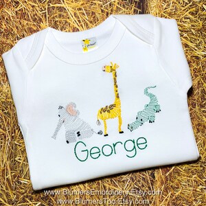 May include: A white short-sleeved baby bodysuit featuring an embroidered design of a gray elephant, a yellow giraffe, and a green alligator. Below the animals, the name George is embroidered in green thread. A small giraffe logo tag is at the neck.