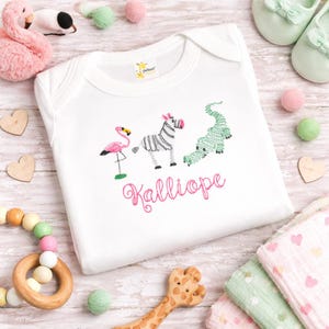 May include: A white short-sleeved baby bodysuit featuring an embroidered design of a pink flamingo, a black and white zebra, and a green alligator. Below the animals, the name Kalliope is embroidered in pink cursive script lettering.