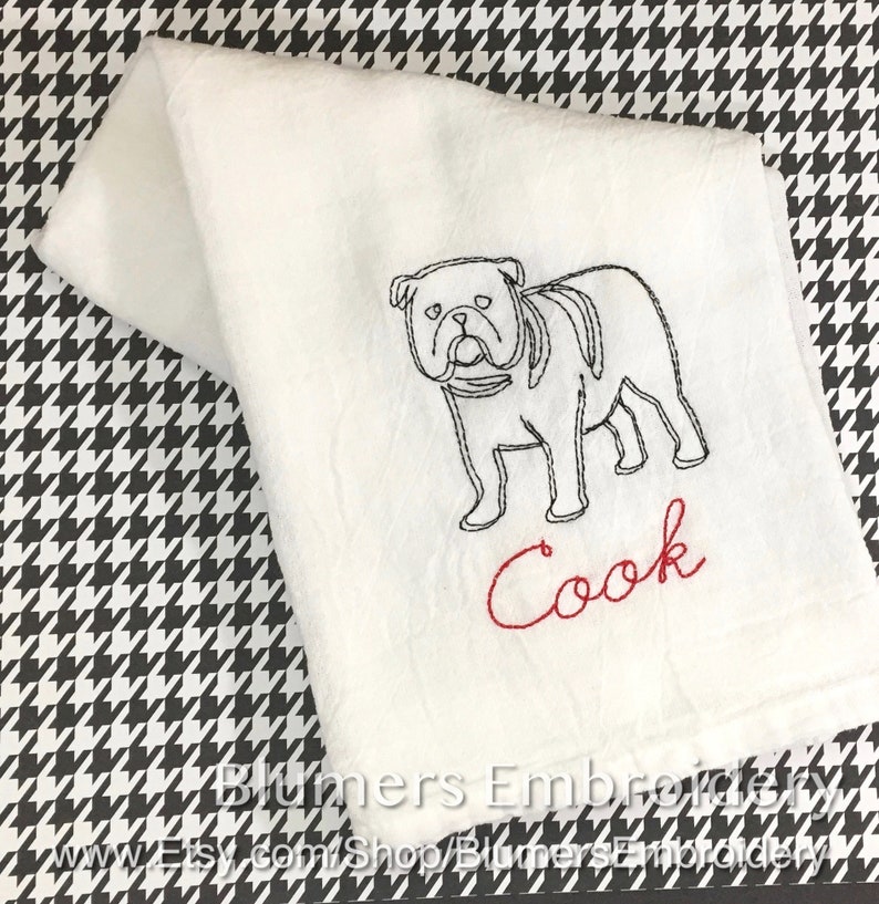 K&ouml;nnte beinhalten: Ein wei&szlig;es K&uuml;chentuch mit einer schwarzen Stickerei, die den Umriss einer Bulldogge im Profil zeigt. Unter dem Hund ist das Wort Cook in roter Schreibschrift gestickt. Das Tuch ist gefaltet und zeigt ein schlichtes, klares Design.
