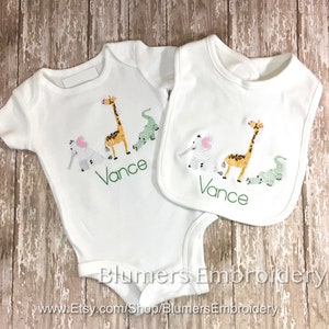 Personalized Jungle Zoo Bodysuit Embroidered Animals T Shirt Burp Cloth Bib Boys Girl Babies Monogrammed Lion Tiger Alligator Giraffe Zebra: Adorable baby bodysuit and bib set featuring embroidered safari animals: giraffe, elephant, and crocodile. The set is perfect for a baby shower gift or to welcome a new baby.  The image shows the items laid out on a rustic wooden background.  The brand name 'Blumers Embroidery' and website address are visible.