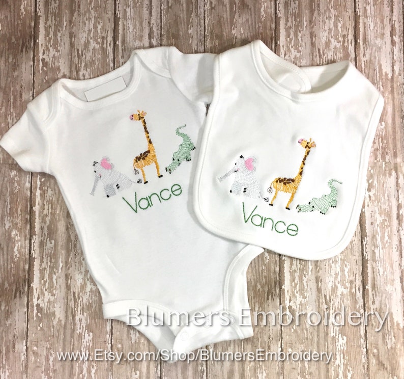 Personalized Jungle Zoo Bodysuit Embroidered Animals T Shirt Burp Cloth Bib Boys Girl Babies Monogrammed Lion Tiger Alligator Giraffe Zebra: Adorable baby bodysuit and bib set featuring embroidered safari animals: giraffe, elephant, and crocodile. The set is perfect for a baby shower gift or to welcome a new baby.  The image shows the items laid out on a rustic wooden background.  The brand name 'Blumers Embroidery' and website address are visible.