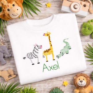 May include: A white sweatshirt featuring an embroidered design of a black and white zebra, a yellow and brown giraffe, and a green crocodile. Below the animals, the name Axel is embroidered in green thread in a simple, clean font.