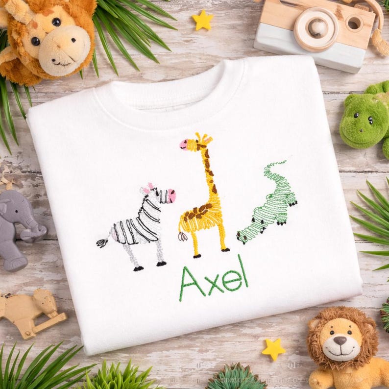 May include: A white sweatshirt featuring an embroidered design of a black and white zebra, a yellow and brown giraffe, and a green crocodile. Below the animals, the name Axel is embroidered in green thread in a simple, clean font.