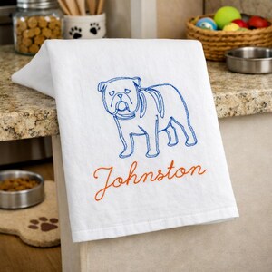 K&ouml;nnte beinhalten: Ein wei&szlig;es K&uuml;chentuch mit einer gestickten blauen Linienzeichnung eines Bulldogs. Unter dem Hund ist der Name Johnston in orangefarbener Schreibschrift gestickt. Das Tuch liegt &uuml;ber der Kante einer K&uuml;chentheke.