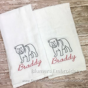 K&ouml;nnte beinhalten: Zwei wei&szlig;e rechteckige K&uuml;chent&uuml;cher mit einer schwarzen Stickerei, die den Umriss einer Bulldogge zeigt. Unter jeder Bulldogge ist der Name Braddy in rotem Schreibschriftgarn gestickt. Der Text Blumers Embroidery und eine URL sind sichtbar.