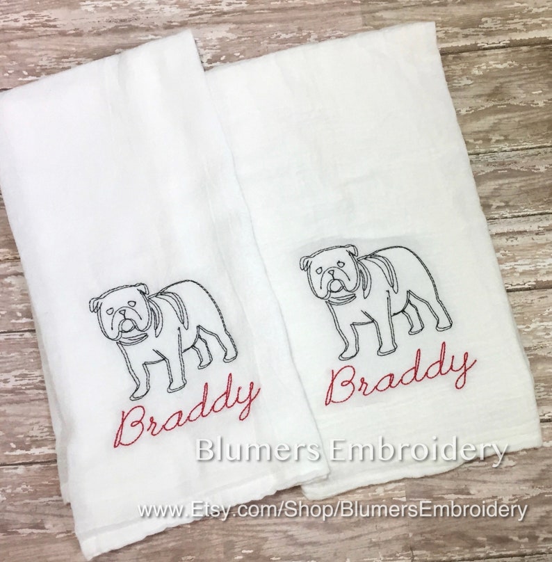 K&ouml;nnte beinhalten: Zwei wei&szlig;e rechteckige K&uuml;chent&uuml;cher mit einer schwarzen Stickerei, die den Umriss einer Bulldogge zeigt. Unter jeder Bulldogge ist der Name Braddy in rotem Schreibschriftgarn gestickt. Der Text Blumers Embroidery und eine URL sind sichtbar.