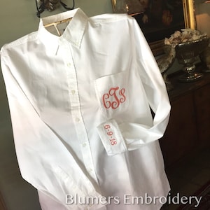 May include: A white long-sleeve button-down shirt with a pocket on the left side. The pocket has a red embroidered monogram "GJS". The cuff of the shirt has a red embroidered date "6-9-18".