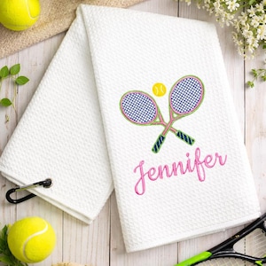 May include: White tennis towel with embroidered crossed rackets, a tennis ball, and the name "Jennifer" in pink script. The towel has a textured pattern and a black carabiner clip. Tennis balls and a tennis racket are in the background.